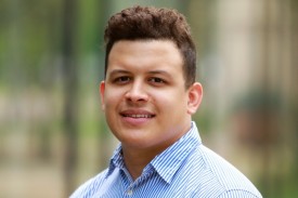 Researcher with dark hair sits outside in a light blue striped button down shirt